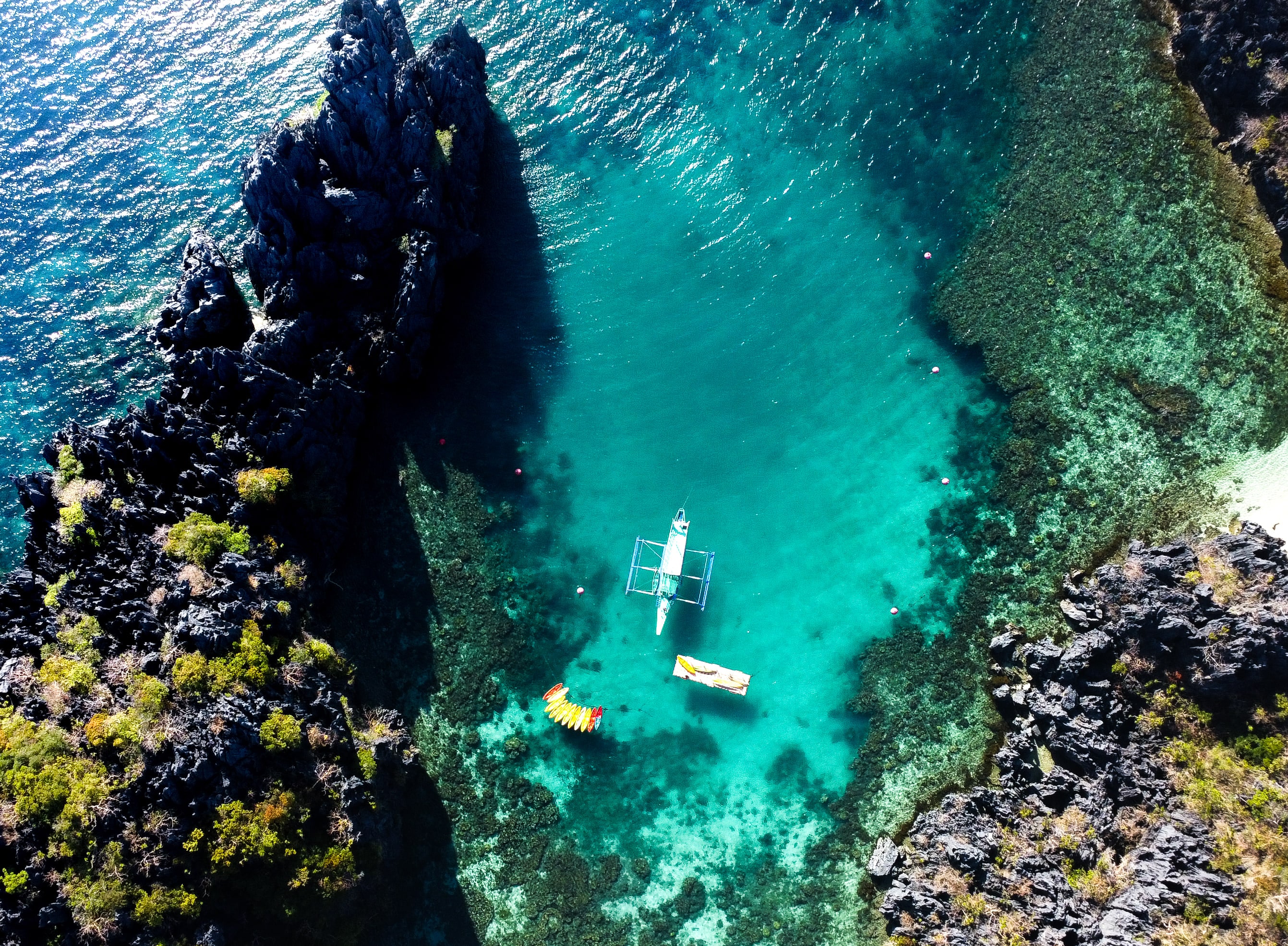 Vista en picado de una laguna resguardada de aguas turquesas transparentes, entre acantilados calizos negros y dentados, donde una barca con balancín, una pequeña barca blanca y un kayak amarillo flotan sobre un arrecife visible.