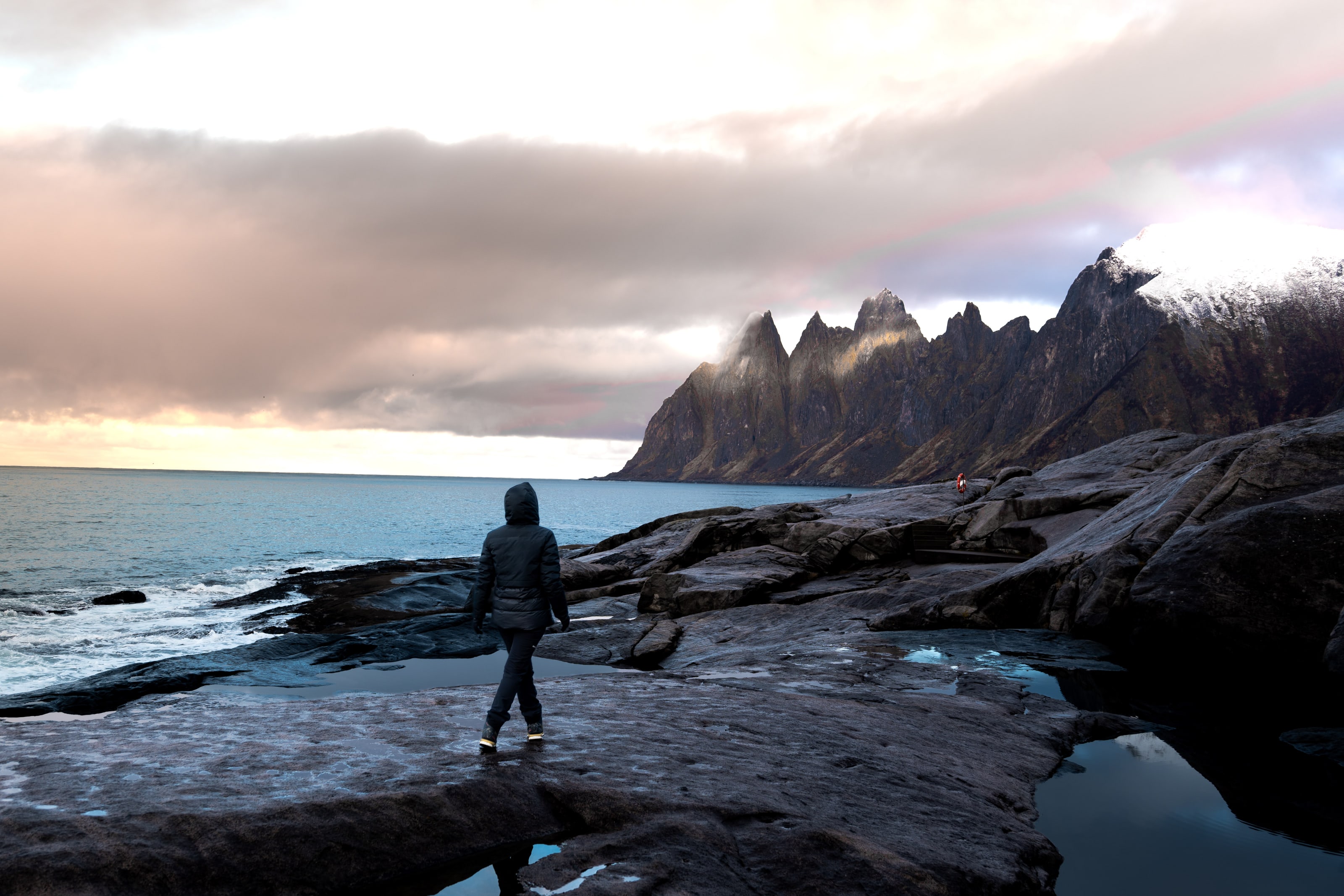 Una figura solitaria, con abrigo oscuro con capucha, está de pie sobre una amplia plataforma de roca mojada, frente a una hilera de picos dentados al otro lado de un mar gris, bajo un banco de nubes bajas encendido por el ocaso.