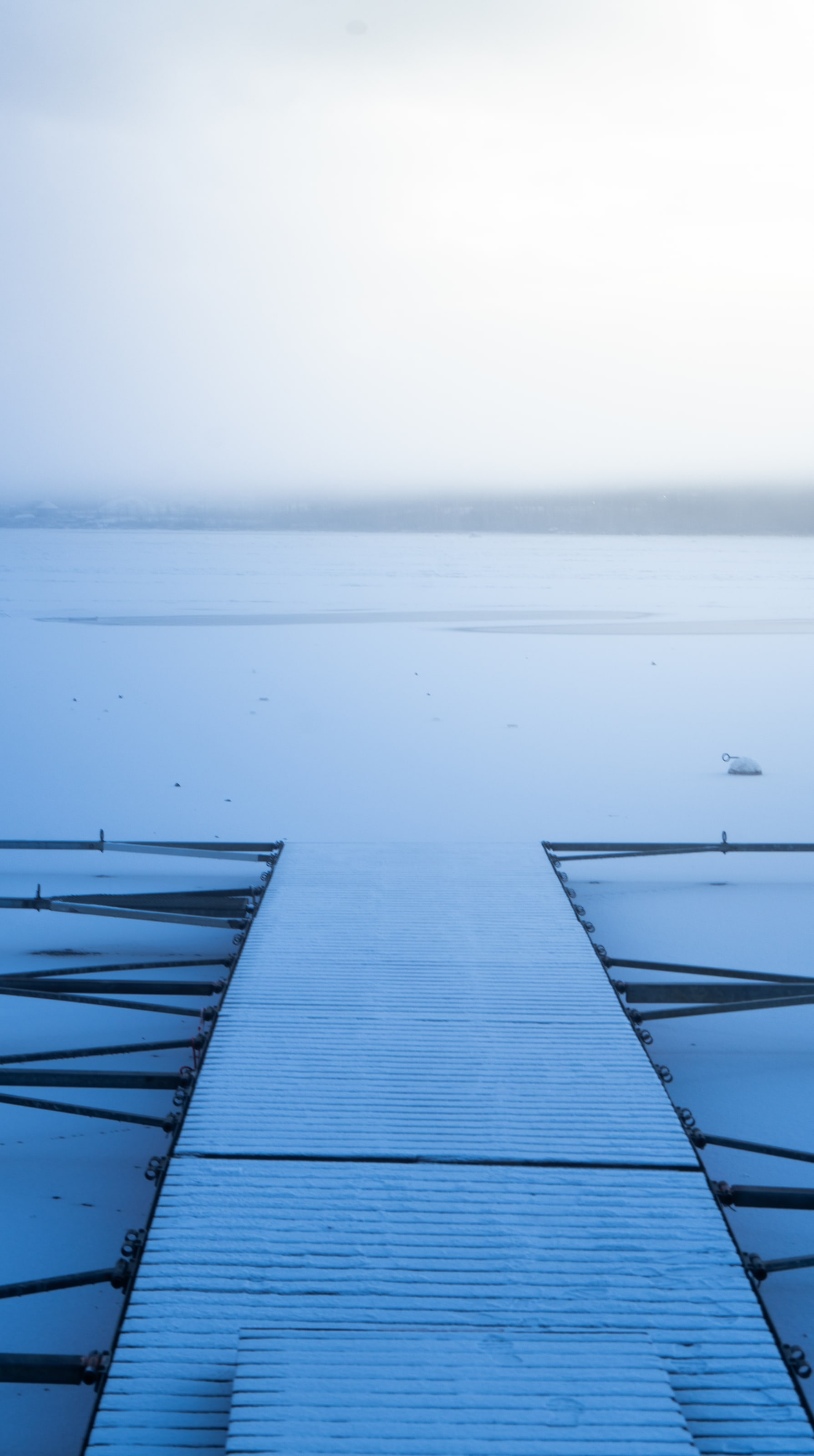 Un muelle estrecho de madera se aleja en línea recta sobre un lago helado espolvoreado de nieve, con su extremo disolviéndose en una niebla blanca donde debería estar la orilla.