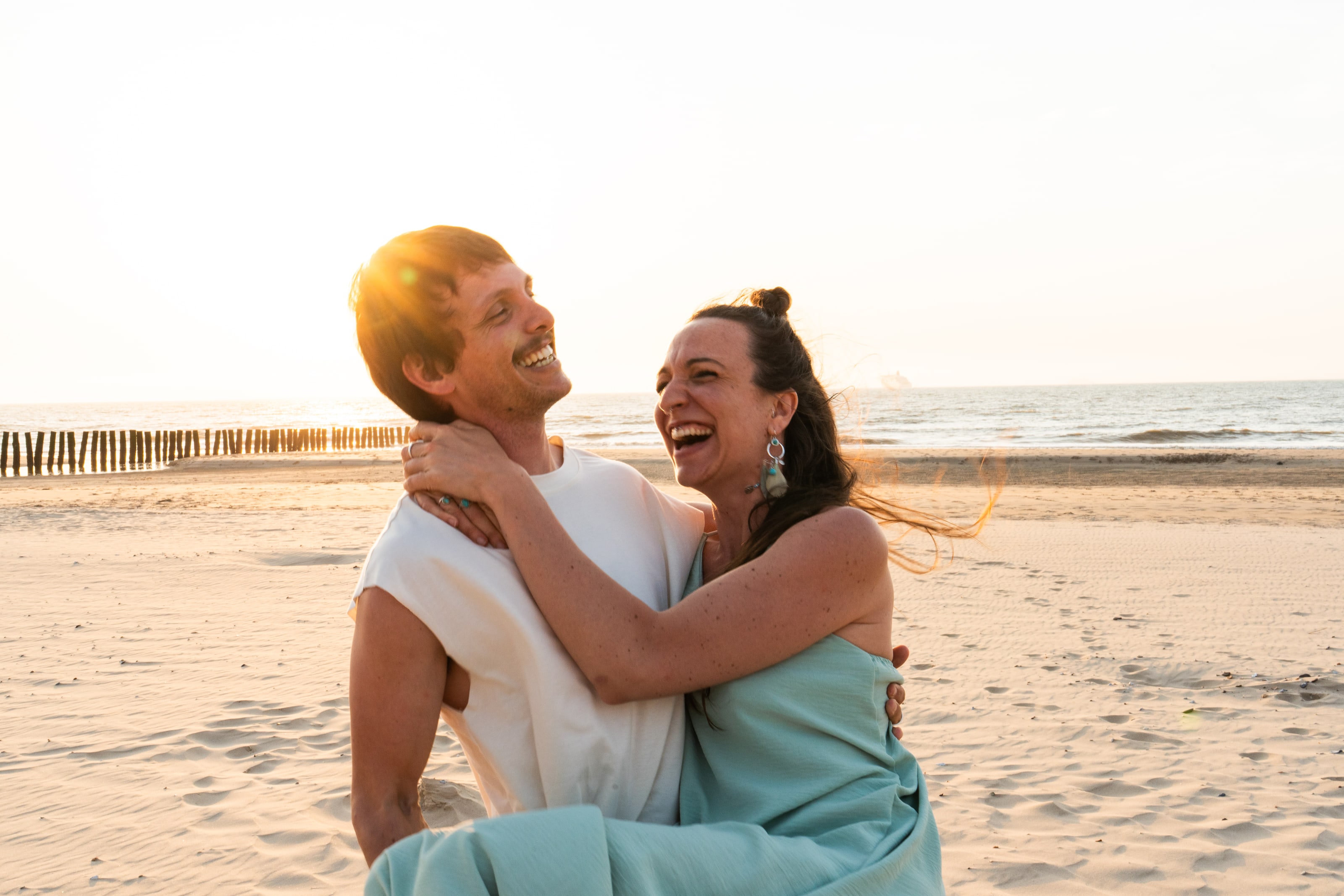 Sur une large plage de sable au coucher du soleil, une femme, les bras autour du cou d'un homme, rit la tête rejetée en arrière, ses cheveux balayés en travers du cadre, le soleil bas éclatant derrière lui.