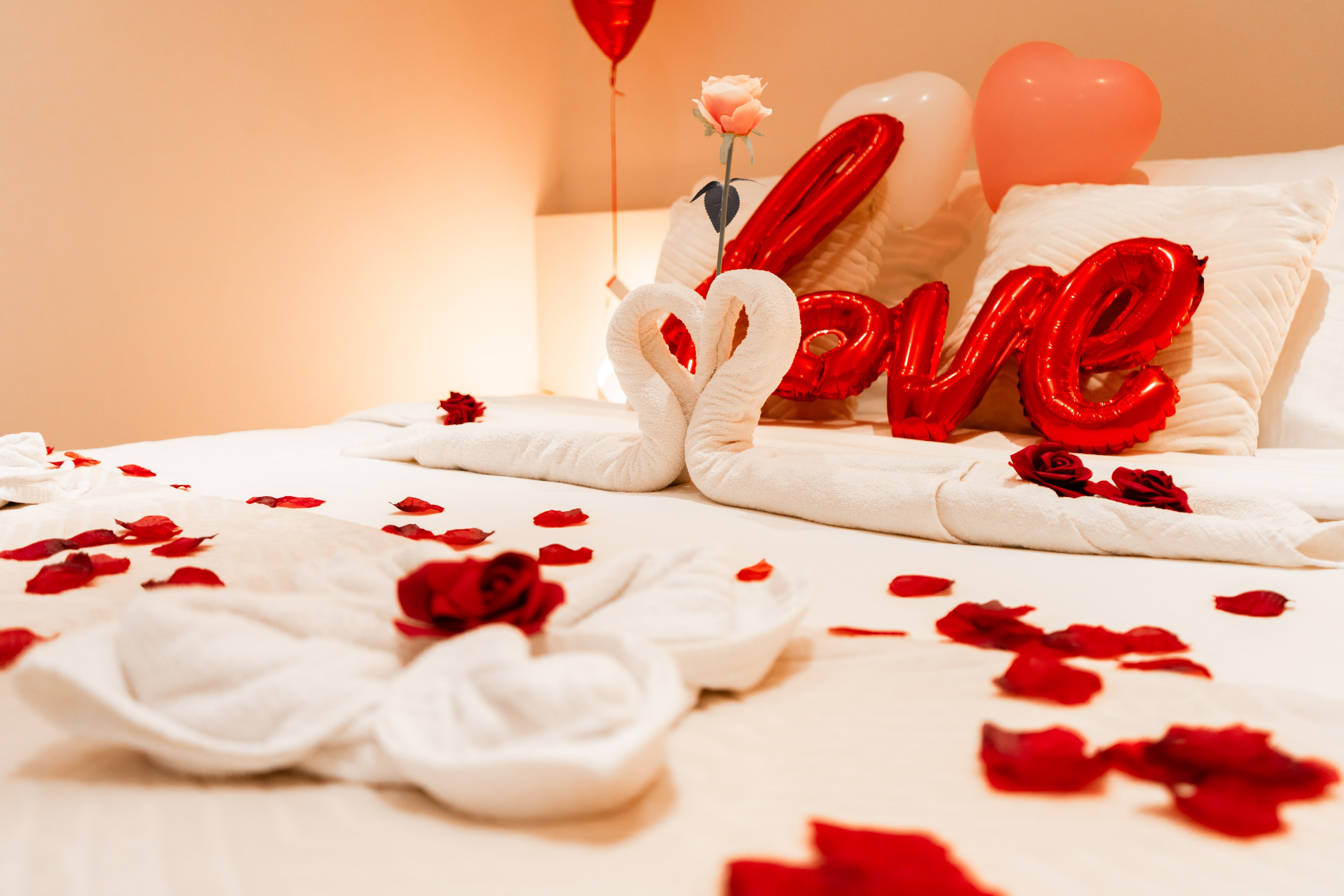 Una cama de matrimonio hecha en blanco, con dos toallas dobladas en forma de cisne que se encuentran formando un corazón, una rosa pálida en un jarrón estrecho detrás, globos de aluminio rojo que forman la palabra «love» apoyados en las almohadas, globos con forma de corazón rojos, blancos y rosados por encima, y pétalos de rosa rojo oscuro esparcidos sobre la colcha y hasta el primer plano.