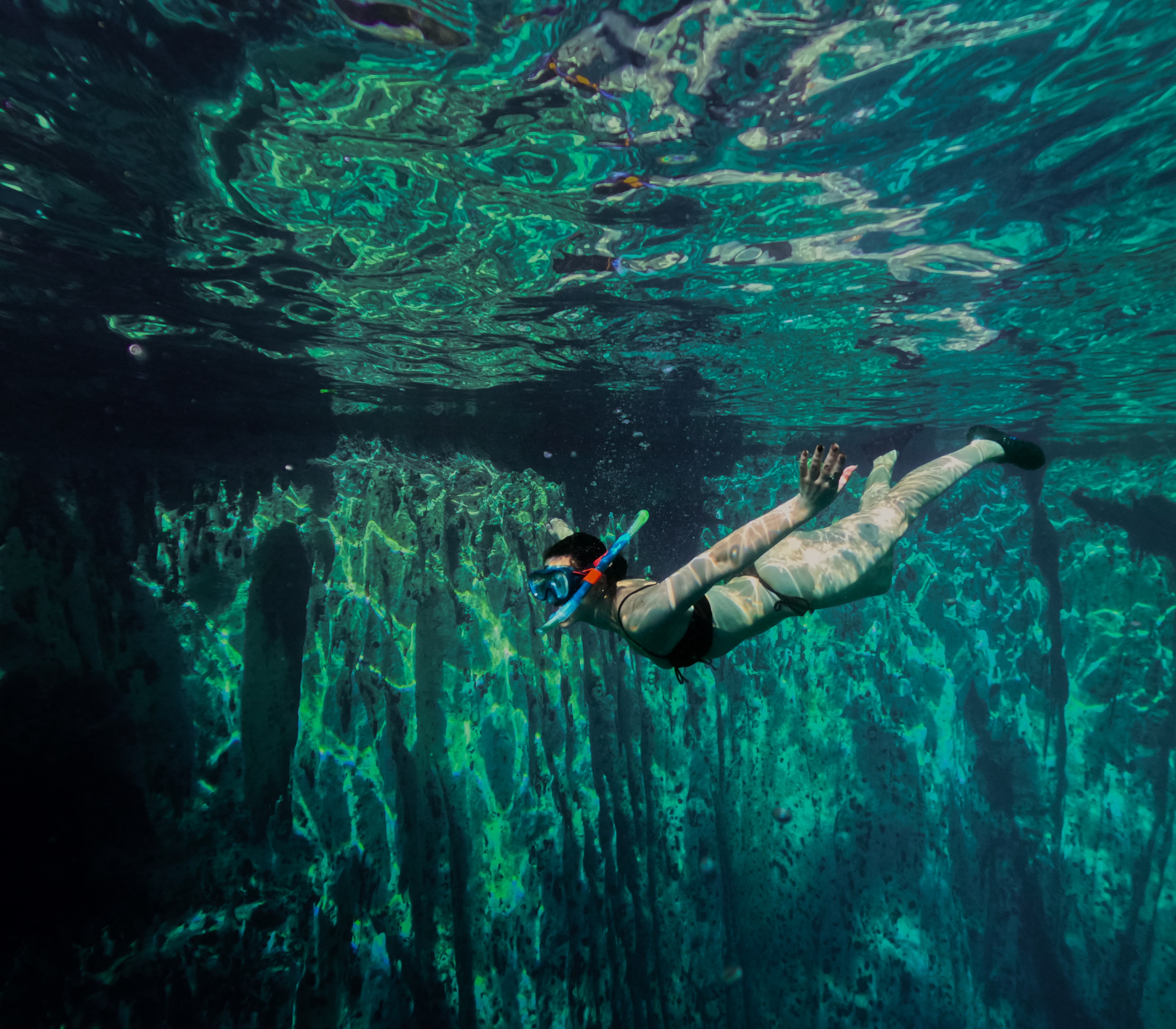 Fotografiado bajo el agua, alguien con máscara y aletas se desliza justo debajo de la superficie, que hace de espejo, sobre una pared caliza estriada que cae hacia una oscuridad verde.