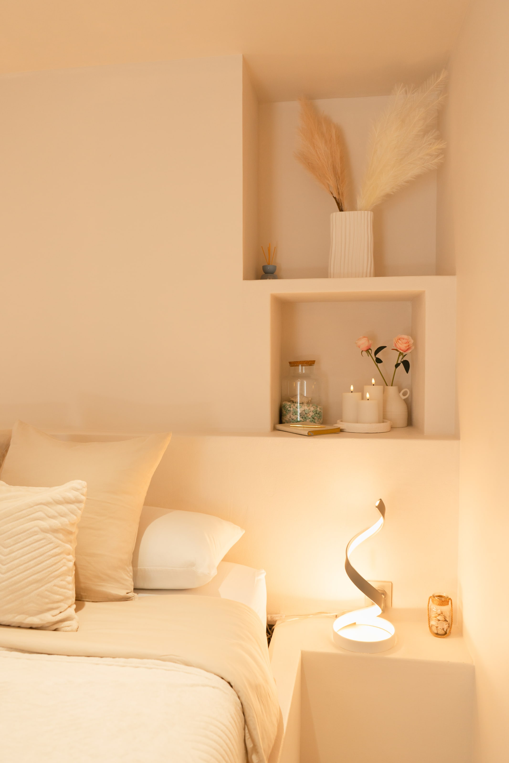 Le coin d'un lit dans une chambre en enduit crème, surmonté de deux niches : l'une avec des herbes de la pampa dans un vase blanc côtelé, l'autre avec des bougies, deux roses roses dans un pichet et un petit livre — et, sur la tablette en dessous, une lampe sculpturale enroulée qui projette une lumière chaude sur le mur.