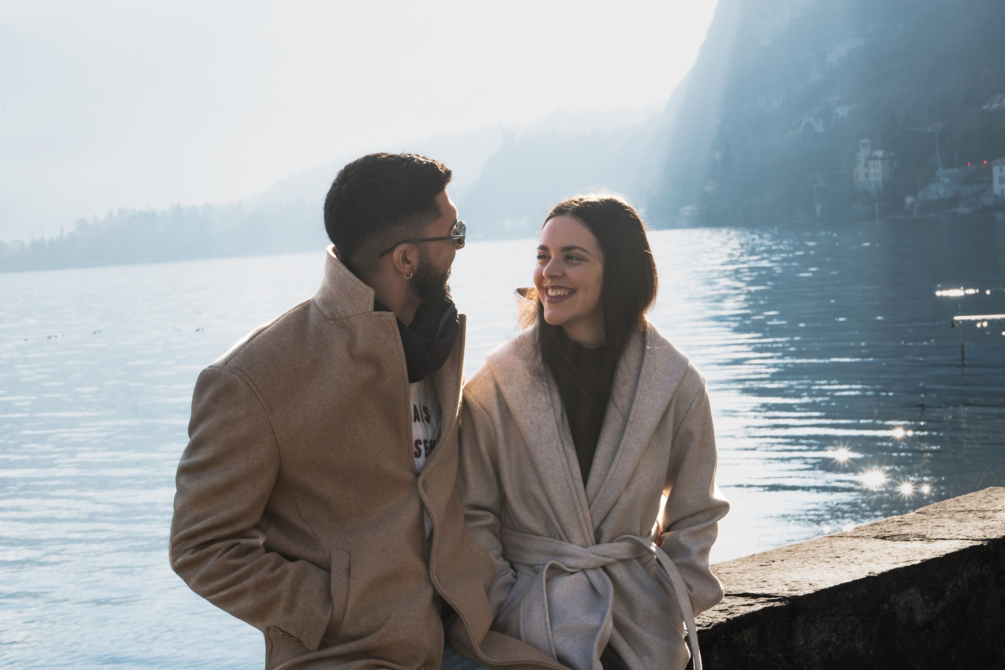 Una pareja con abrigos de lana claros se sienta en un muro de piedra al borde de un lago, vueltos el uno hacia el otro entre risas, con el sol bajo estallando en puntos de luz sobre el agua a su espalda.